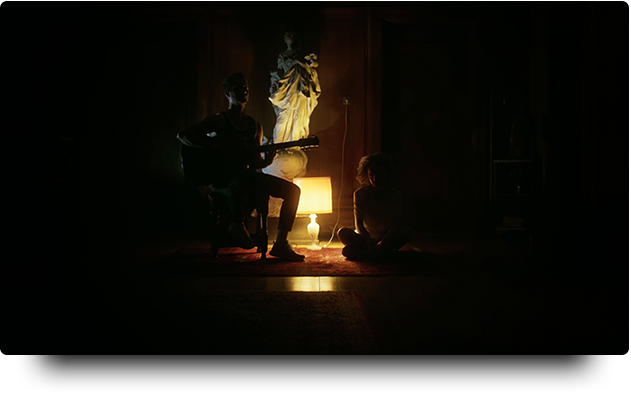 eine steinbüste wird vom gelben licht einer einzelnen alten schirmlampe beleuchtet. davor sitzen zwei musiker namens tomsis